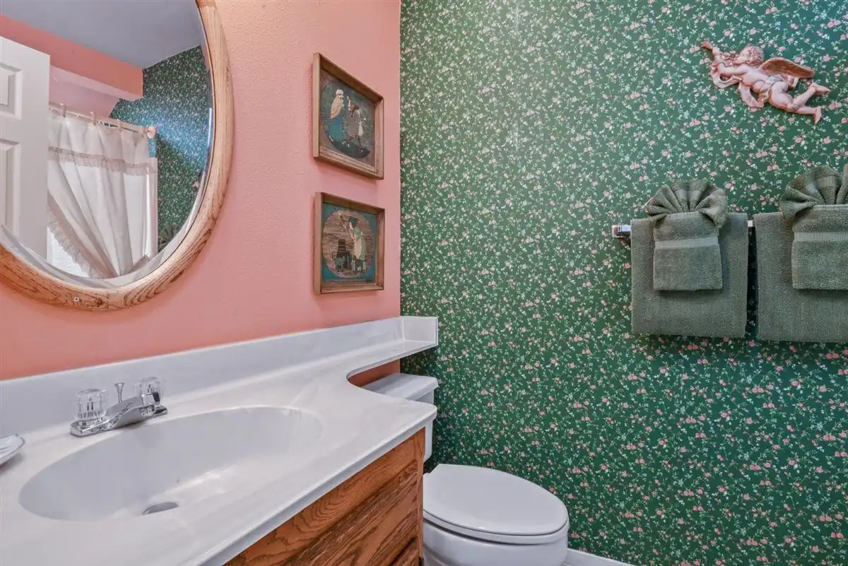 A wood-framed oval mirror and two vintage pictures above a sink, next to plush green towels on floral wallpaper.