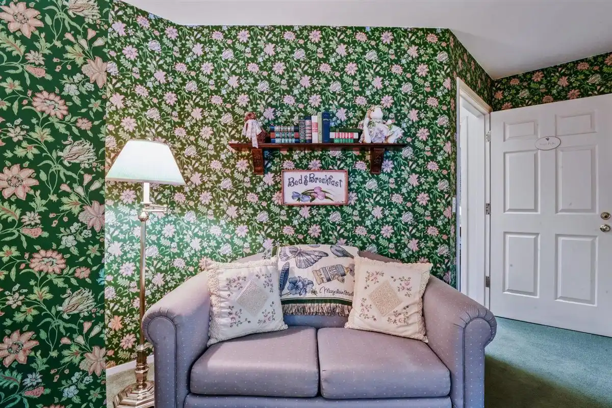 Loveseat with a lamp under a bookshelf against a floral green wallpaper.